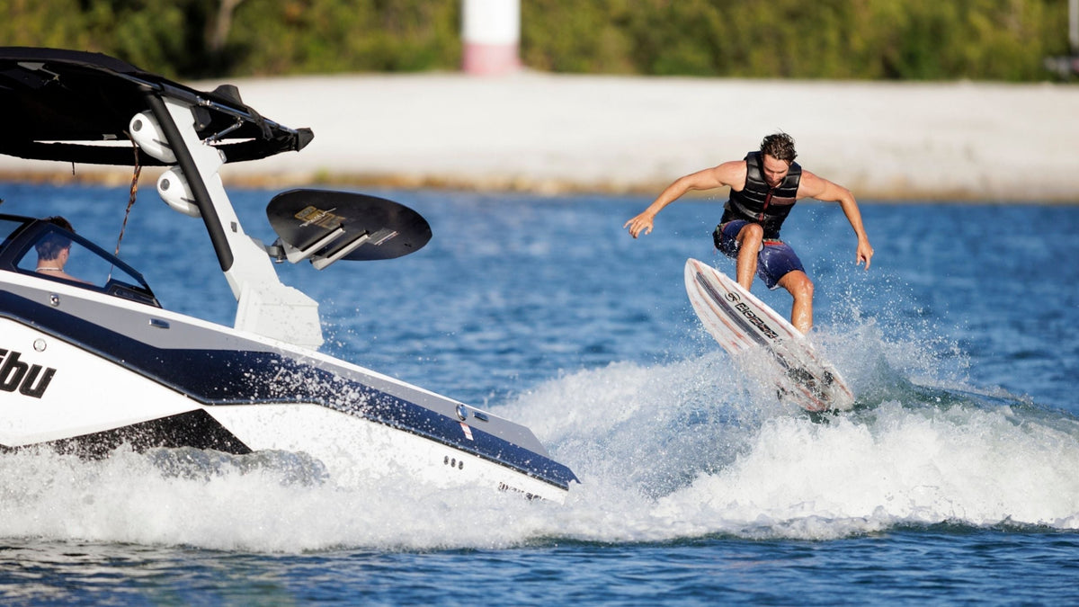 A guy wakesurfing behind a Malibu boat doing an ollie off the wake on the new 2026 Ronix Atlantik Wakesurf Board.
