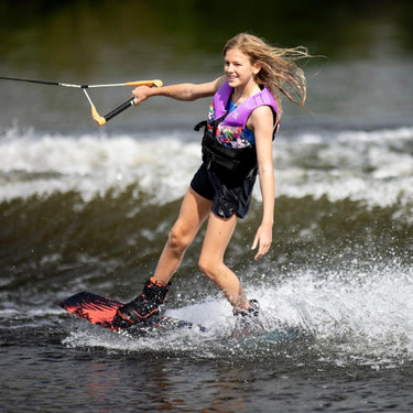 Young girl grom riding with one hand off the handle and cruising comfortably outside the wake on the 2026 Ronix Krush 125cm wakeboard setup.