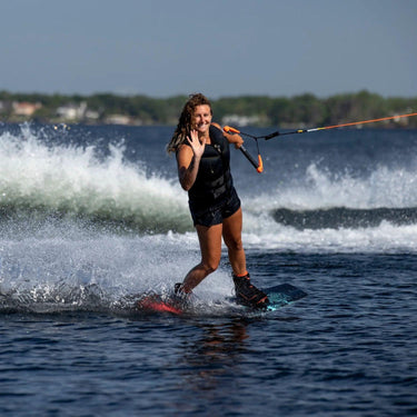 Progressing girl wakeboarder cruising with one hand off the handle and waving on the 2026 Ronix Krush 125cm wakeboard package.