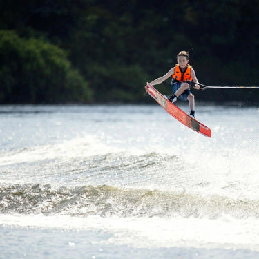 Young grom wakeboarder confidently launching off the wake and locking in a clean tail grab on the complete 2026 Ronix Kid's District 129cm wakeboard package.