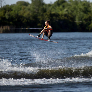 Young grom wakeboarder confidently executing a clean wake-to-wake jump and grab on the complete 2026 Ronix Kid's District 129cm wakeboard package.