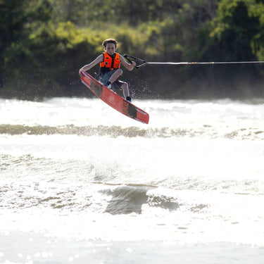 Young youth rider launching off the wake and locking in a stylish tail grab on the 2026 Ronix Kid's District 129cm wakeboard setup